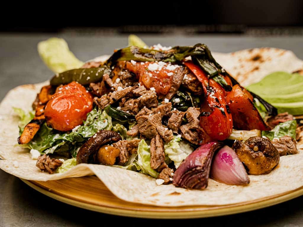 Flame grilled meats and vegetables (red bell and jalapeno peppers, red and green onions, tomato, sweet potato & mushrooms) on a bed of romaine in a creamy cilantro dressing atop a charred tortilla with re-fried beans, avocado, roasted tomato salsa and cotija cheese.